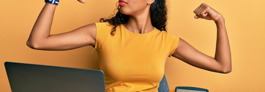 rethinking employee benefits: a professionally-dressed business woman flexing her biceps while sitting at her laptop.