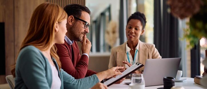 An insurance broker reviews a policy document with a couple at their home, explaining the terms on a clipboard while the man and woman look on thoughtfully over coffee.